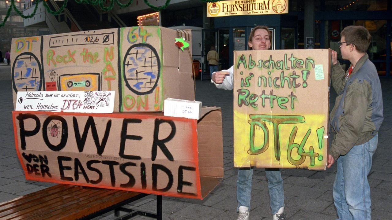 Zu den vielen Aktionen für den Erhalt des Jugendradios DT64 zählte auch eine Demonstration mit Mahnwache auf dem Berliner Alexanderplatz am 17.12.1991. Zwei Jugendliche stehen vor bunt bemalten Radios aus Pappe 