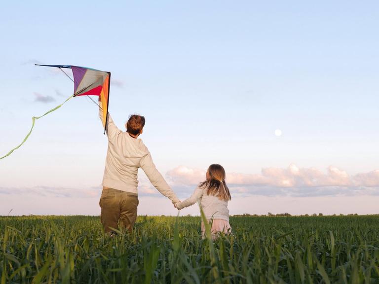 Ein Mann hält einen Drachen, während er mit seiner Tochter an der Hand durch ein Feld läuft.