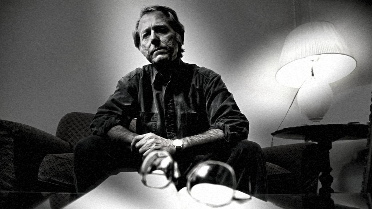 Schwarzweißaufnahme von Don DeLillo, der in einer dramatisierenden Perspektive vor einem Buch sitzt. Schwarzweißaufnahme von Don DeLillo, der in einer dramatisierenden Perspektive vor einem Buch sitzt.