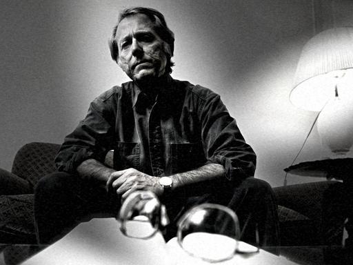 Schwarzweißaufnahme von Don DeLillo, der in einer dramatisierenden Perspektive vor einem Buch sitzt.