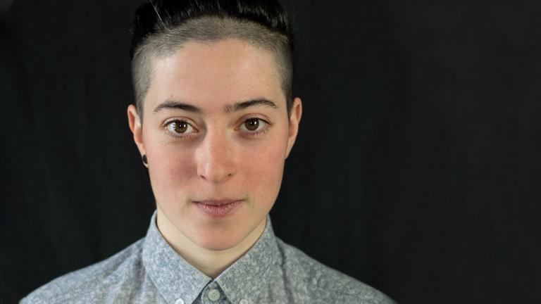Inna Barinberg steht vor einem schwarzen Hintergrund, trägt ein graues Hemd und schaut freundlich in die Kamera. Ihre Haare sind hochgesteckt und teilweise abrasiert. Inna Barinberg steht vor einem schwarzen Hintergrund, trägt ein graues Hemd und schaut freundlich in die Kamera. Ihre Haare sind hochgesteckt und teilweise abrasiert.