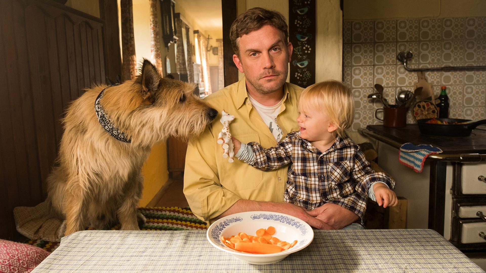 Szene aus dem Film "Leberkäsjunkie": Dorfpolizist Franz Eberhofer (Sebastian Bezzel) sitzt in seiner Küche am Esstisch, auf dem Schoß ein kleines Kind, daneben ein Hund.