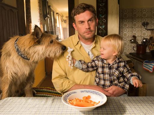 Szene aus dem Film "Leberkäsjunkie": Dorfpolizist Franz Eberhofer (Sebastian Bezzel) sitzt in seiner Küche am Esstisch, auf dem Schoß ein kleines Kind, daneben ein Hund.