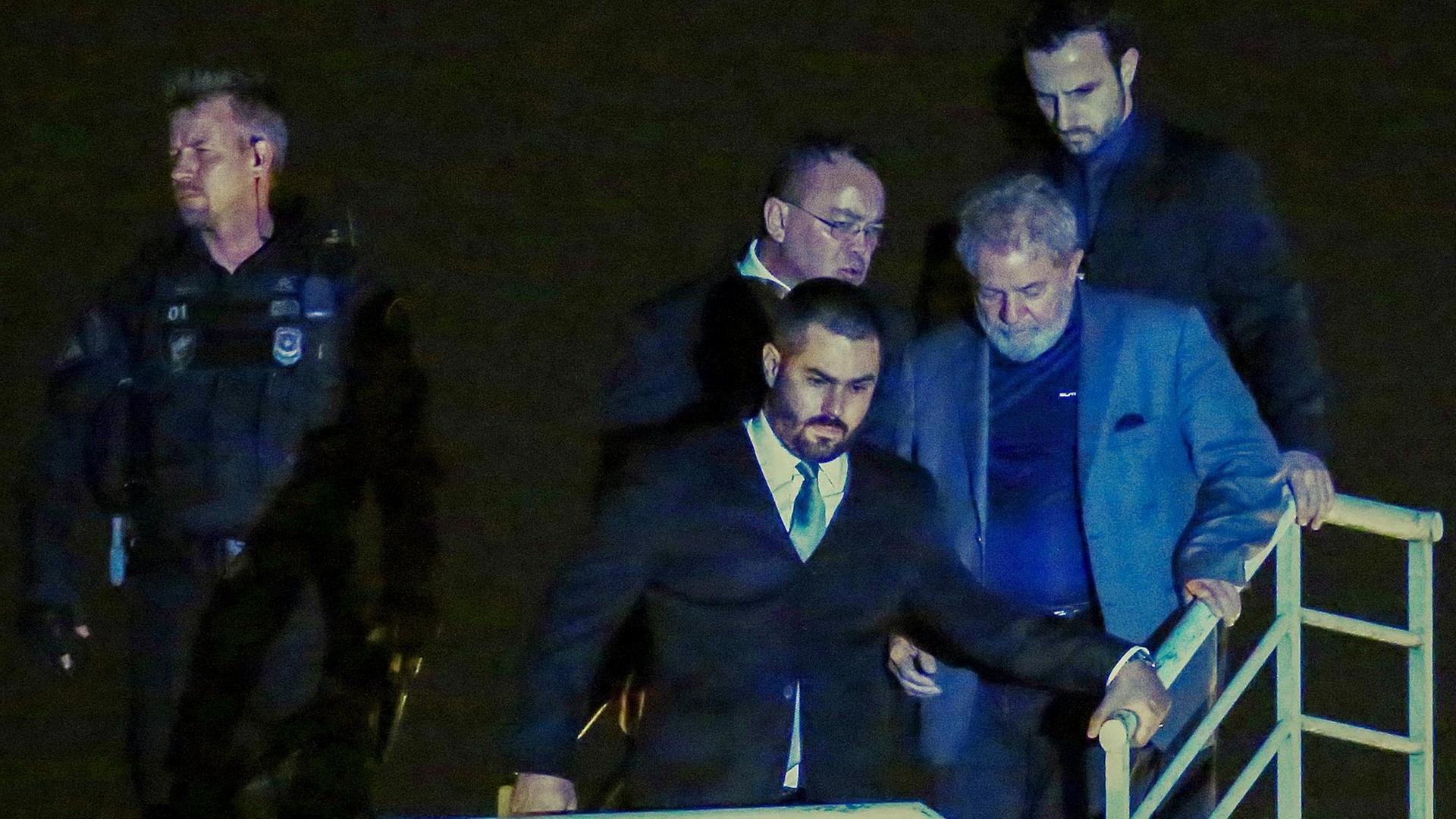 Ein grobkörniges Foto von Lula und mehreren Sicherheitsbeamten, die in der Nachnt ein Geländer herabsteigen. Ein grobkörniges Foto von Lula und mehreren Sicherheitsbeamten, die in der Nachnt ein Geländer herabsteigen.
