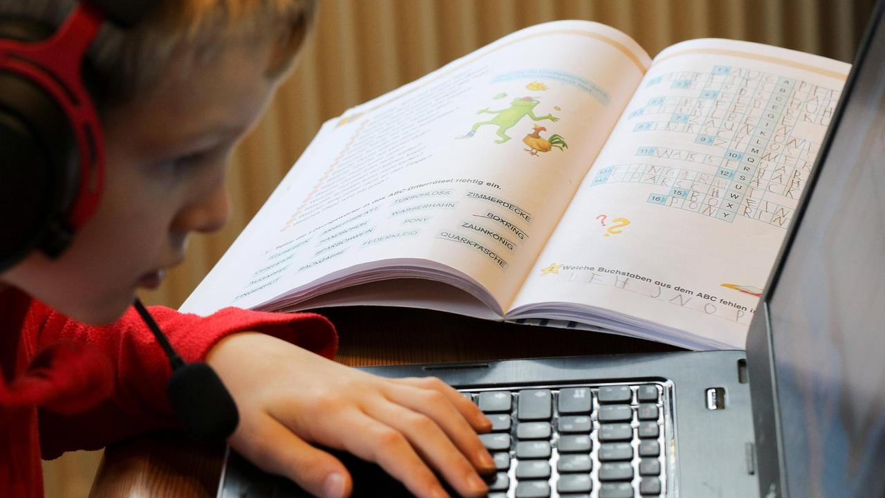 Eine Junge sitzt vor einem aufgeklappten Laptop mit Kopfhörern und Mikrofon und macht Homeschooling. Auf dem Tisch liegen verschiedene Schulbücher. Eine Junge sitzt vor einem aufgeklappten Laptop mit Kopfhörern und Mikrofon und macht Homeschooling. Auf dem Tisch liegen verschiedene Schulbücher.