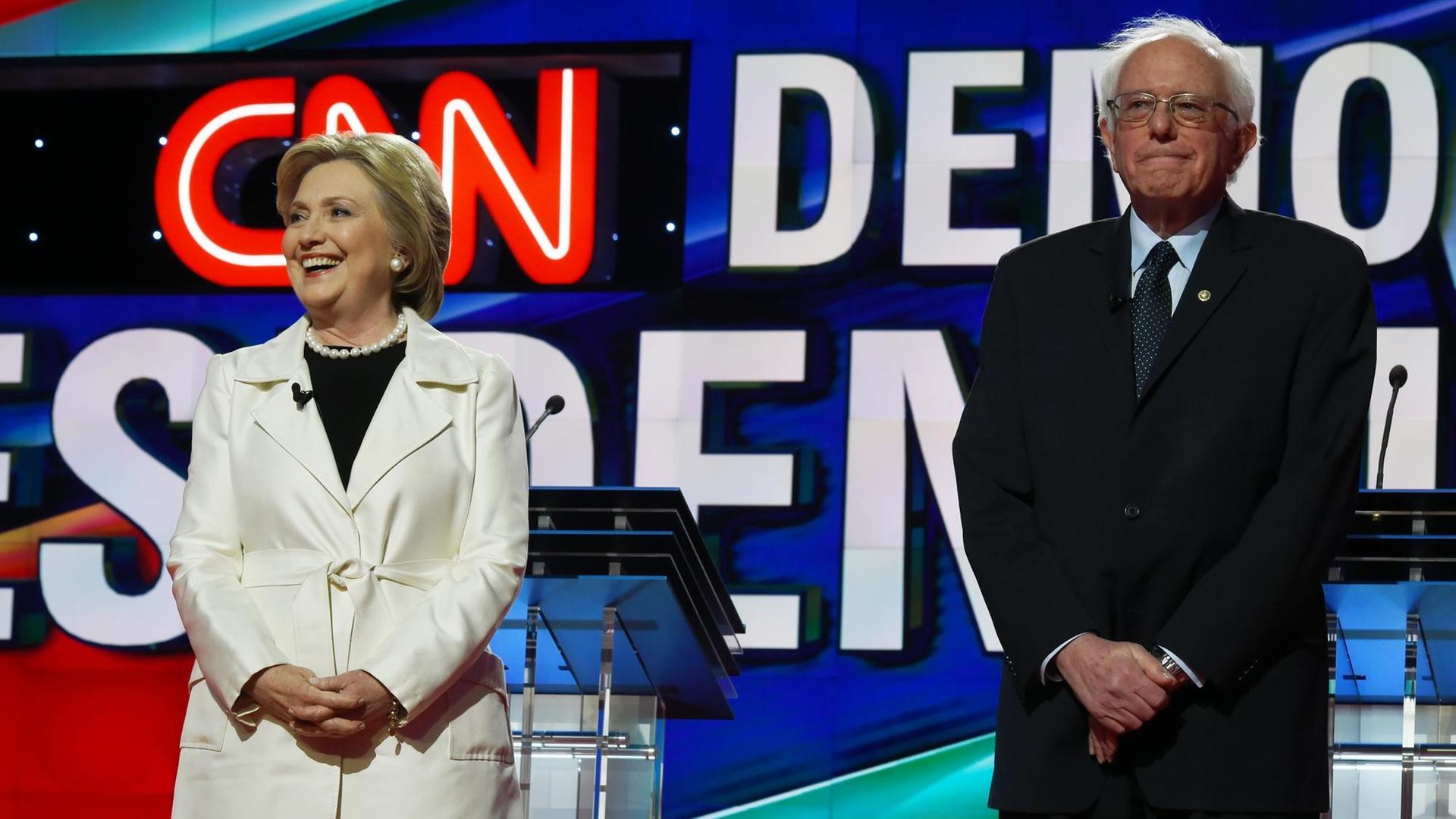 Hillary Clinton und Bernie Sanders stehen nebeneinander auf einer CNN-Bühne. Clinton lacht, Sanders schaut grimmig.