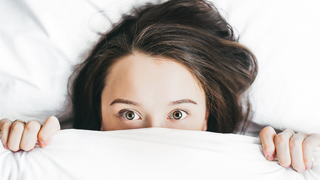 Eine Frau liegt im Bett und zieht angsterfüllt die Bettdecke bis unter die Augen.