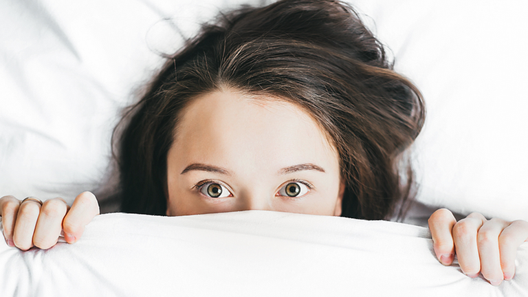Eine Frau liegt im Bett und zieht angsterfüllt die Bettdecke bis unter die Augen. Eine Frau liegt im Bett und zieht angsterfüllt die Bettdecke bis unter die Augen.