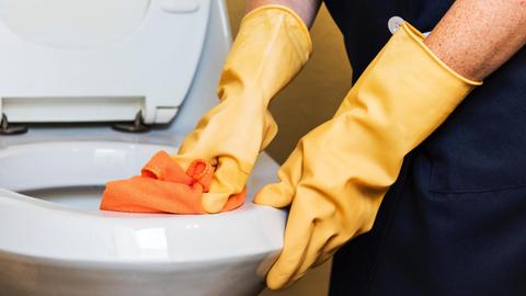 Hände in gelben Handschuhen putzen mit einem orangenen Tuch ein Klo.