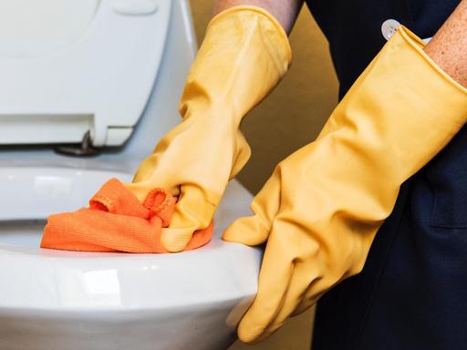 Hände in gelben Handschuhen putzen mit einem orangenen Tuch ein Klo.
