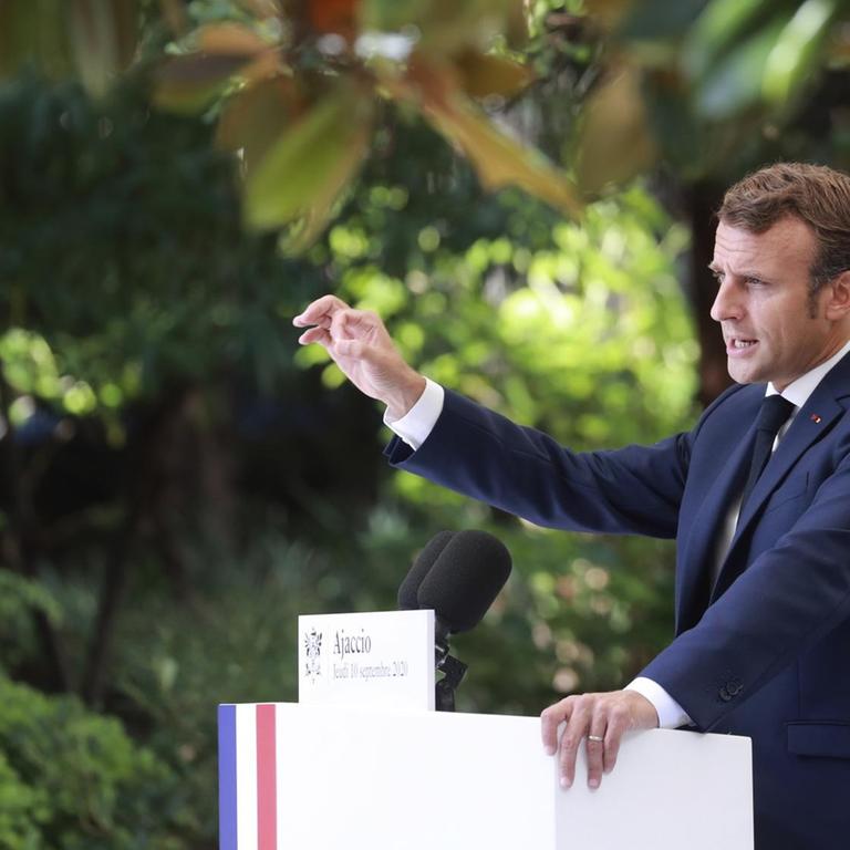 Macron steht unter Bäumen an einem Rednerpult mit den französischen Farben, spricht in ein Mikrofon und gestikuliert mit der erhobenen Hand. Er ist von der Seite fotografiert.