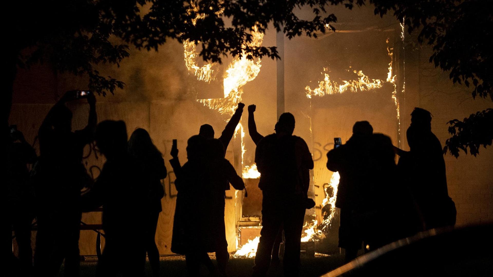 Menschen als Silhouetten vor einem brennenden Geschäft, das im Zuge der Unruhen in Brand gesteckt wurde. Menschen als Silhouetten vor einem brennenden Geschäft, das im Zuge der Unruhen in Brand gesteckt wurde.
