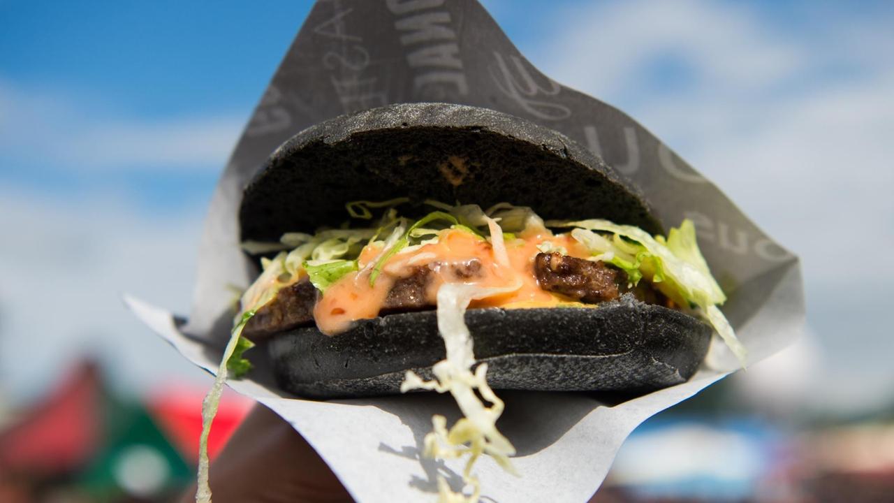 Eine Hand hält einen schwarzen Burger in die Luft. Dahinter blauer Himmel. Eine Hand hält einen schwarzen Burger in die Luft. Dahinter blauer Himmel.