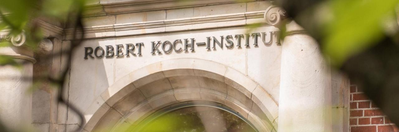Der Eingang des Robert Koch-Instituts ist mit seinem Namenslogo durch Bäume hindurch zu sehen. Der Eingang des Robert Koch-Instituts ist mit seinem Namenslogo durch Bäume hindurch zu sehen.