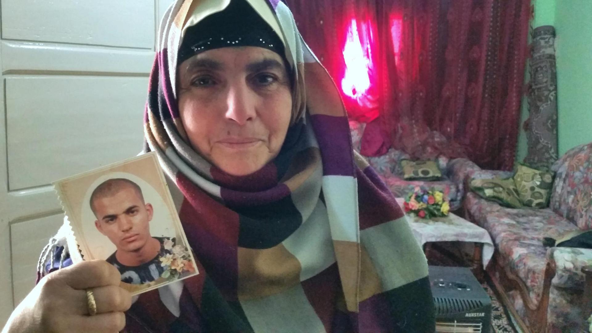 Eine Mutter in Tunesien zeigt das Bild ihres Sohnes in die Kamera. Sie steht im Wohnzimmer.