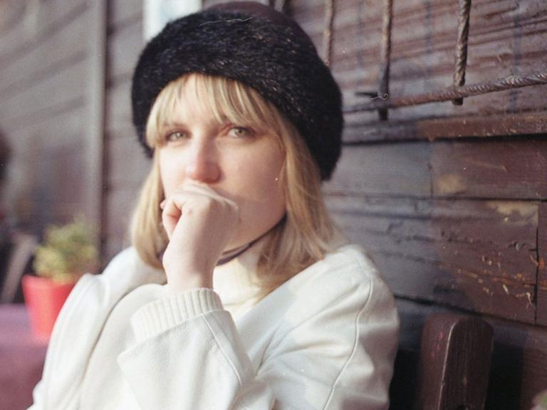 Frieda Paris sitzt auf einem Stuhl in einem Gartencafé, hat eine dunkle Mütze auf und einen weißen Mantel an Frieda Paris sitzt auf einem Stuhl in einem Gartencafé, hat eine dunkle Mütze auf und einen weißen Mantel an