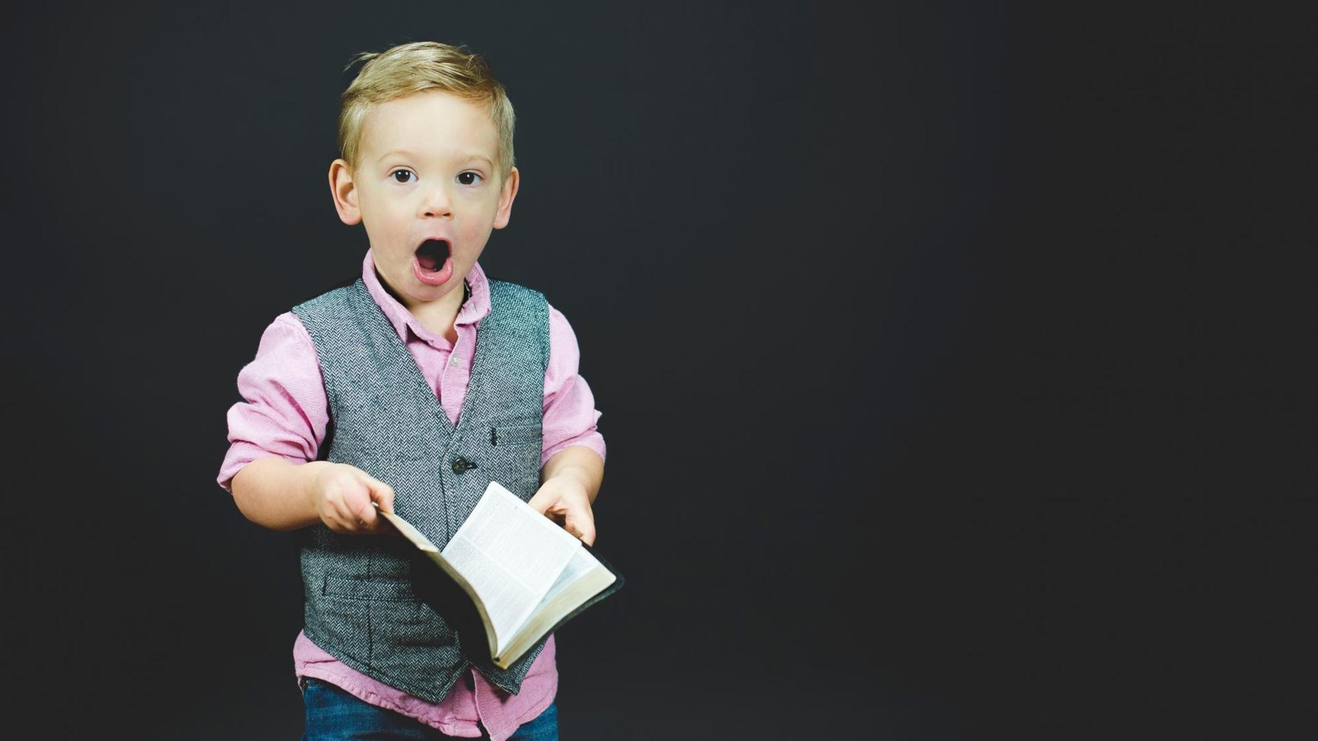 Ein Junge in grauer Weste und pinkem Hemd hält mit großen Augen und offenem Mund ein aufgeschlagenes Buch in Händen. Ein Junge in grauer Weste und pinkem Hemd hält mit großen Augen und offenem Mund ein aufgeschlagenes Buch in Händen.