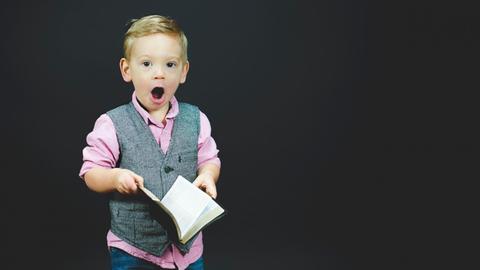 Ein Junge in grauer Weste und pinkem Hemd hält mit großen Augen und offenem Mund ein aufgeschlagenes Buch in Händen.