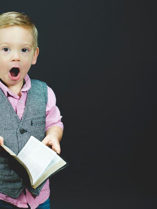 Ein Junge in grauer Weste und pinkem Hemd hält mit großen Augen und offenem Mund ein aufgeschlagenes Buch in Händen.