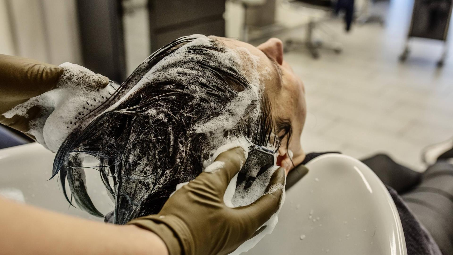 Eine Frau sitzt im Frisiersalon und hat den Kopf ins Waschbecken zurückgelegt. Sie hat Schaum im Haar, die behandschuhten Hände des Friseurs sind zu sehen, wie sie der Frau Schaum aus dem Haar spülen. Eine Frau sitzt im Frisiersalon und hat den Kopf ins Waschbecken zurückgelegt. Sie hat Schaum im Haar, die behandschuhten Hände des Friseurs sind zu sehen, wie sie der Frau Schaum aus dem Haar spülen.