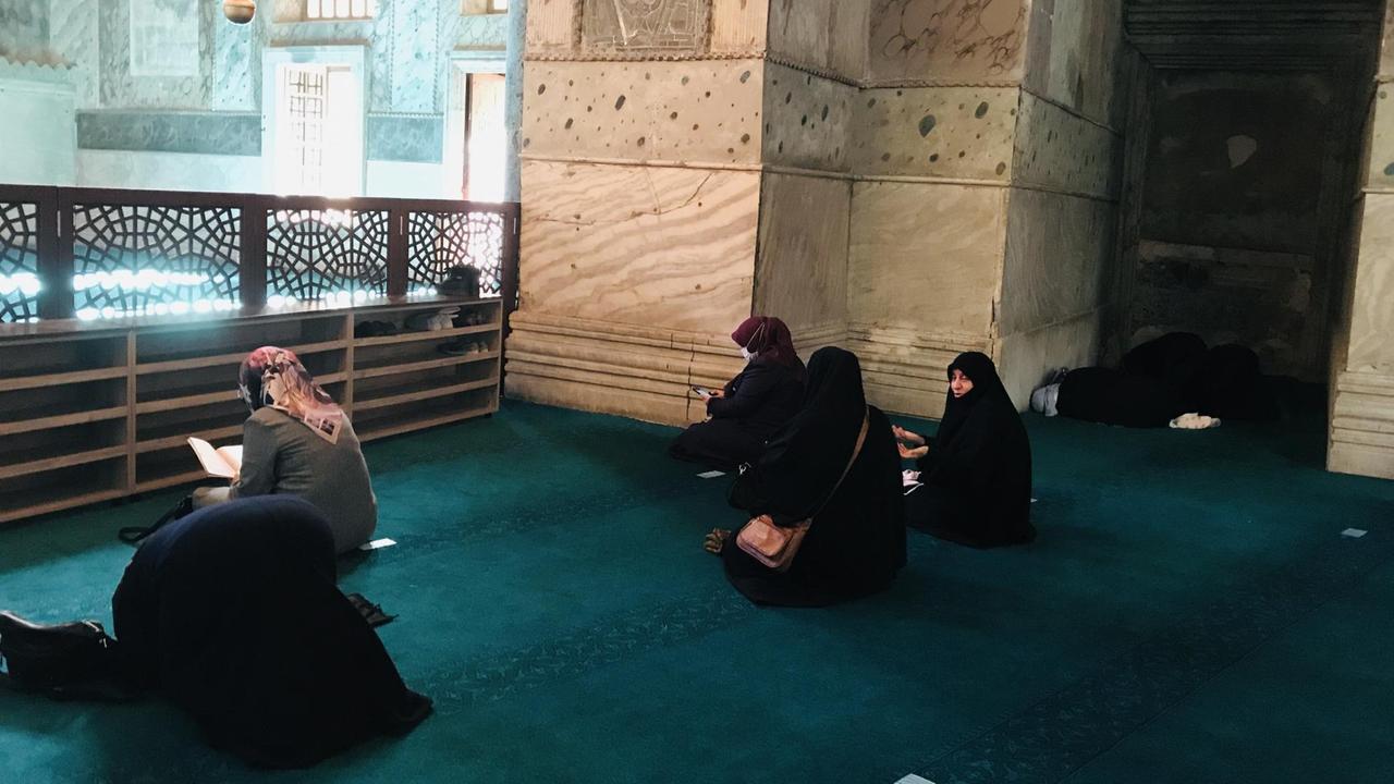 Mehrere Frauen mit schwarzer Kopfbedeckung sitzen auf einem türkisfarbenen Teppich vor einem Altar. Mehrere Frauen mit schwarzer Kopfbedeckung sitzen auf einem türkisfarbenen Teppich vor einem Altar.