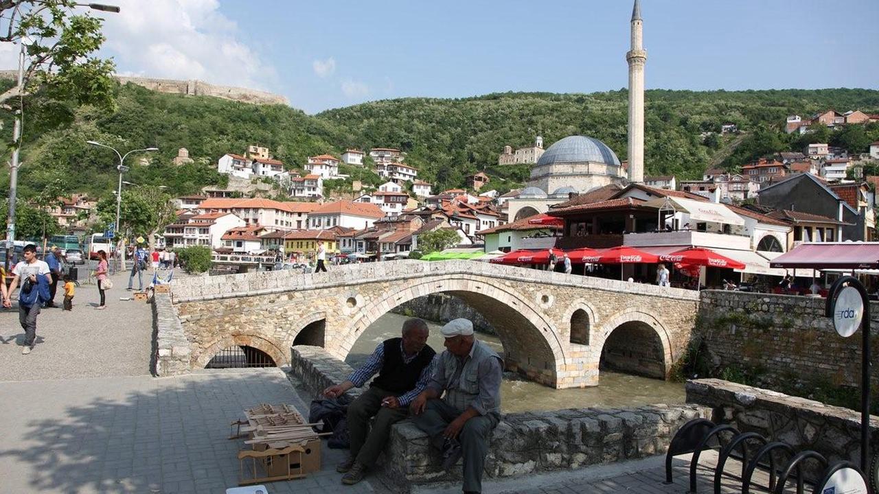 Stadtansicht des kosovarischen Prizren, im Vordergrund eine historische Brücke, dahinter eine Moschee und Wohnhäuser an einem Hügel
