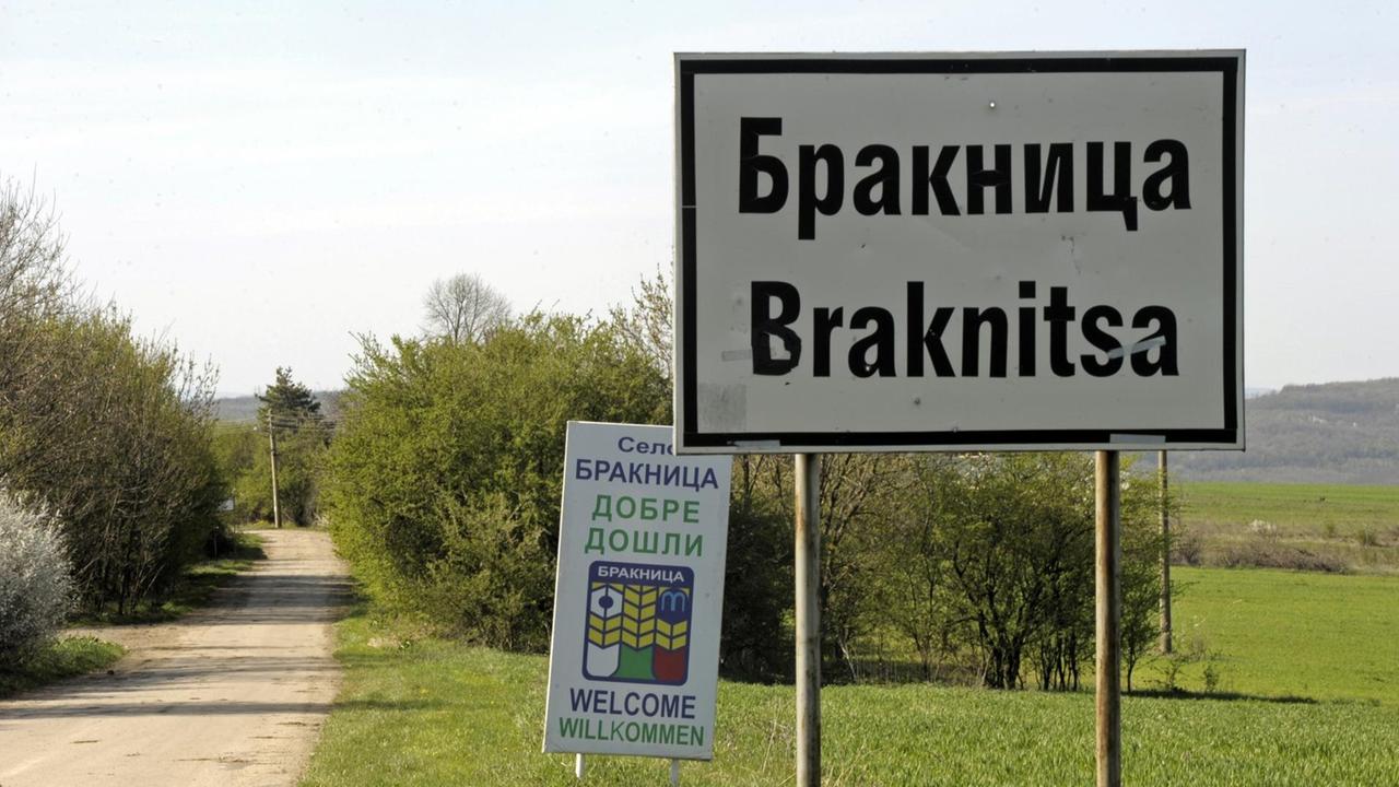Eine Wiese mit einem Stadtschild, auf dem Braknitsa steht. Dahinter ein weiteres Schild mit Willkommensgrüßen in verschiedenen Sprachen. Eine Wiese mit einem Stadtschild, auf dem Braknitsa steht. Dahinter ein weiteres Schild mit Willkommensgrüßen in verschiedenen Sprachen.