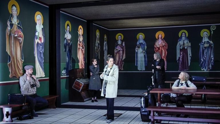Drei Schauspielerinnen und zwei Schauspieler sitzen und stehen in einem Klassenzimmer mit Ikonen an der Wand. Drei Schauspielerinnen und zwei Schauspieler sitzen und stehen in einem Klassenzimmer mit Ikonen an der Wand.