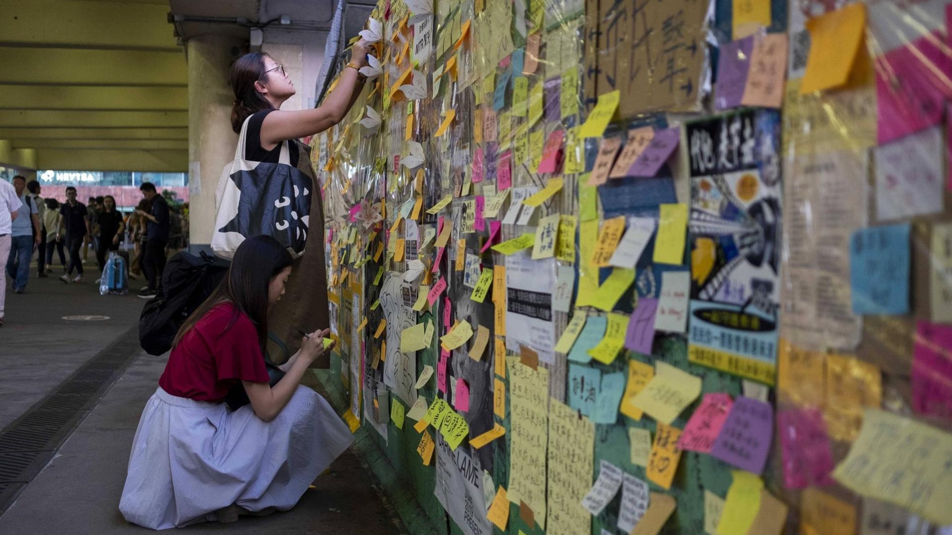 Zwie Frauen schreiben auf Post-its an einer "Lennon Wall" in Hongkong, einer Wand, an der bereits zahlreiche Notizen und Zeichnungen hängen. Zwie Frauen schreiben auf Post-its an einer "Lennon Wall" in Hongkong, einer Wand, an der bereits zahlreiche Notizen und Zeichnungen hängen.