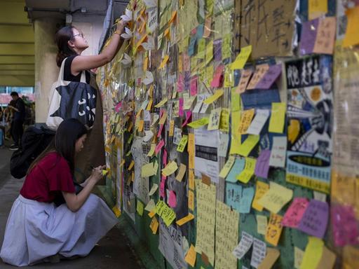 Zwie Frauen schreiben auf Post-its an einer "Lennon Wall" in Hongkong, einer Wand, an der bereits zahlreiche Notizen und Zeichnungen hängen.