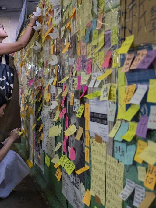 Zwie Frauen schreiben auf Post-its an einer "Lennon Wall" in Hongkong, einer Wand, an der bereits zahlreiche Notizen und Zeichnungen hängen.