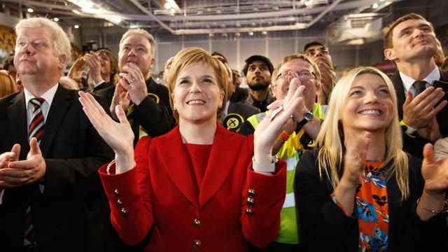 Sturgeon steht in einem roten Kostüm zwischen Parteifreunden, lächelt und klatscht.