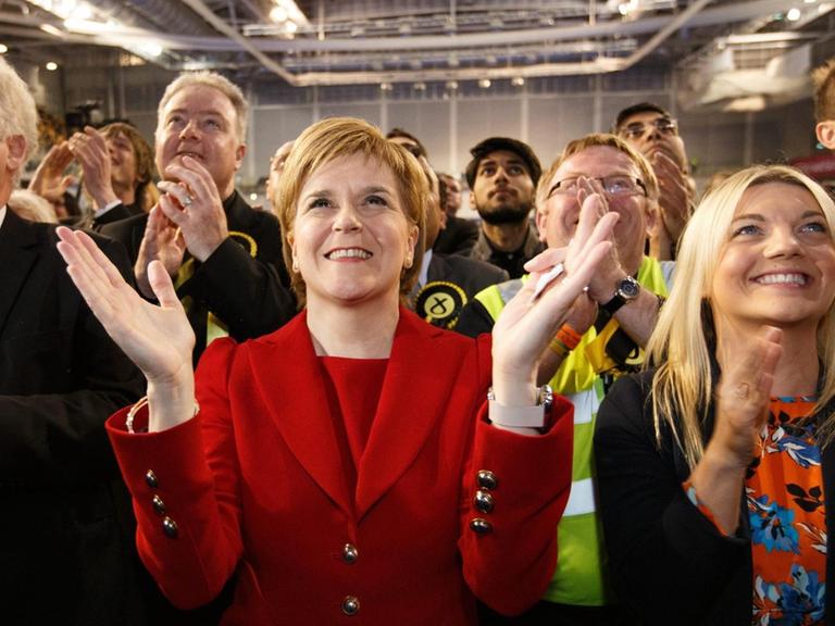 Sturgeon steht in einem roten Kostüm zwischen Parteifreunden, lächelt und klatscht.