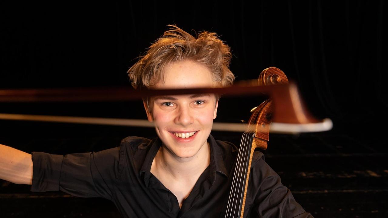 Porträt eines jungen Mannes mit etwas strubbeligen, blondem Haar, der seinen Cellobogen breit lachend in den Vordergrund hält.