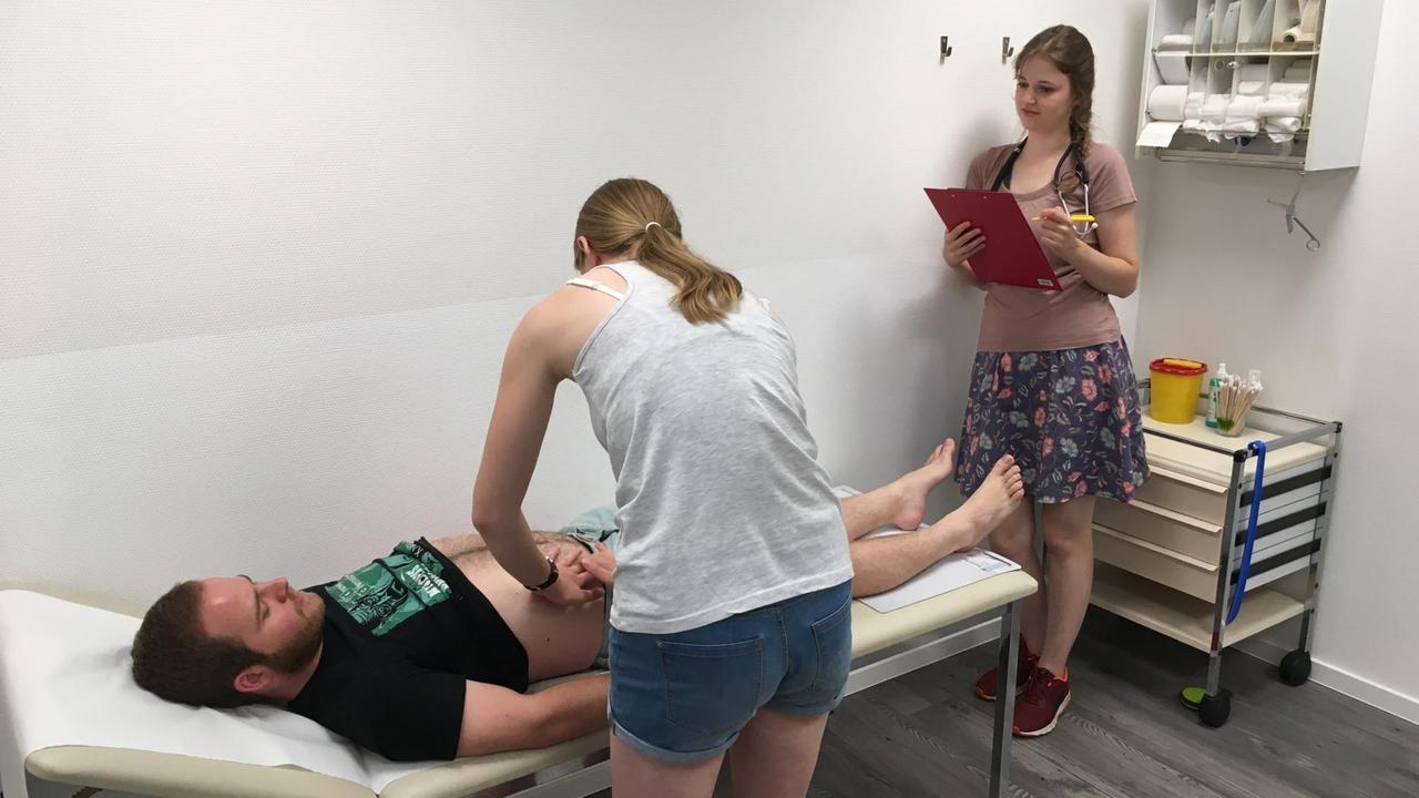 Zwei Medizinstudentinnen üben mit Hilfe eines Schauspielers das Erstuntersuchungsgespräch mit Patienten. Der Patientendarsteller liegt auf einer Liege und hat den Bau frei gemacht, so das eine der Studentinnen ihn abtasten kann. Ihre Studienkollegin steht rechts von ihr mit einem Klemmbrett und macht sich Notizen. Zwei Medizinstudentinnen üben mit Hilfe eines Schauspielers das Erstuntersuchungsgespräch mit Patienten. Der Patientendarsteller liegt auf einer Liege und hat den Bau frei gemacht, so das eine der Studentinnen ihn abtasten kann. Ihre Studienkollegin steht rechts von ihr mit einem Klemmbrett und macht sich Notizen.