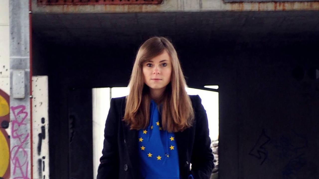 Eine junge Frau mit langen dunklen Haaren in einem alten Industriegebäude: Sie trägt ein blaues Shirt mit Europa-Logo (gelbe Sterne), darüber einen schwarzen Blazer. 