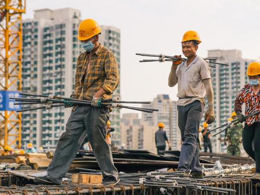 Drei Bauarbeiter mit gelben Helmen tragen Metallstreben auf einer Baustellenkonstruktion.