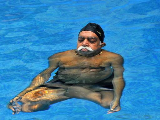 Ein indischer Mann mit weißem Schnurrbart praktiziert Yoga in einem Schwimmbecken.