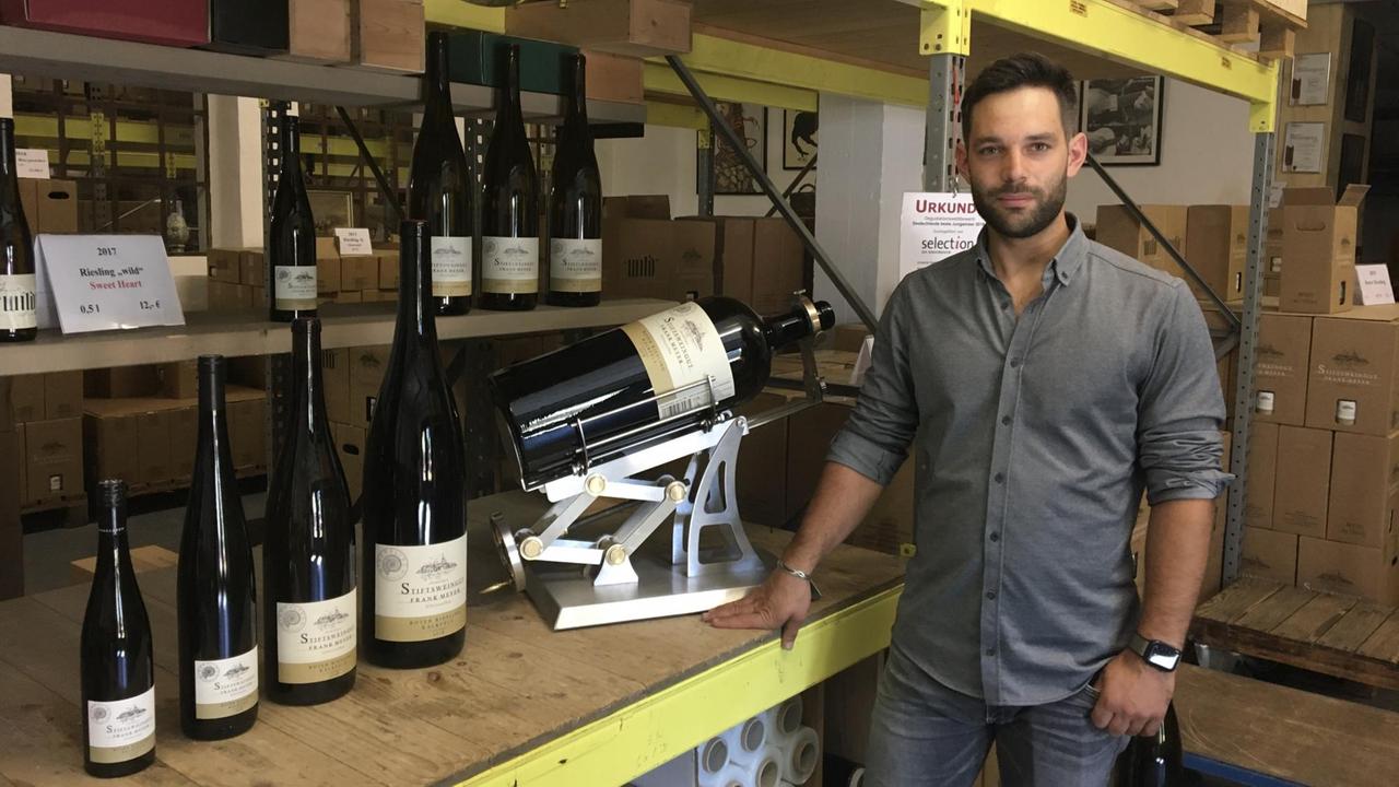 Auf einem Tisch stehen kleine, mittlere und sehr große gefüllt braune Weinflaschen, daneben ein junger Mann mit dunkelbraunen Haaren und dunkelbraunem Bart im grauen Hemd. Auf einem Tisch stehen kleine, mittlere und sehr große gefüllt braune Weinflaschen, daneben ein junger Mann mit dunkelbraunen Haaren und dunkelbraunem Bart im grauen Hemd.
