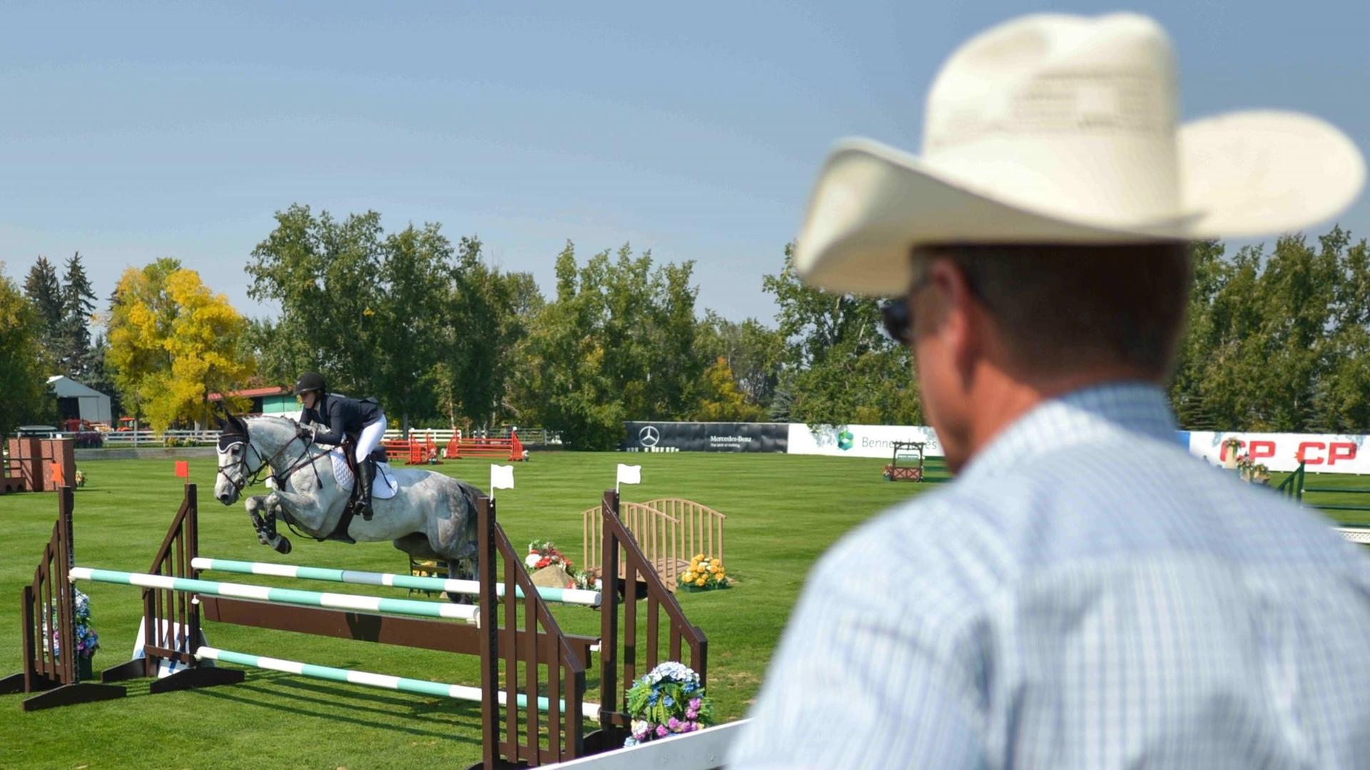 Ein Mann mit Cowboy-Hut beobachtet einen Springreiter des Meadows Cup. Ein Mann mit Cowboy-Hut beobachtet einen Springreiter des Meadows Cup.