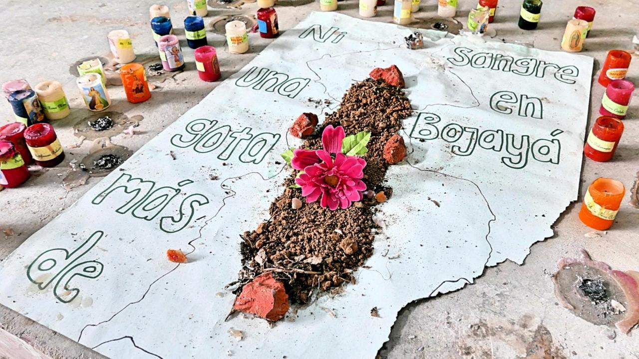 Ein weißes Plakat liegt auf dem Boden, ist von Kerzen umgeben. In der Mitte des Plakates liegt etwas Erde und ein paar Blumen und darauf steht: "Kein Tropfen Blut mehr in Bojayá".
