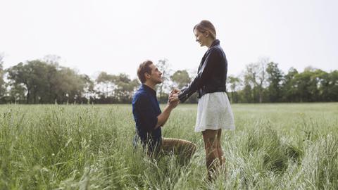 Ein Mann kniet auf einer Wiese vor einer jungen Frau und macht ihr einen Heiratsantrag