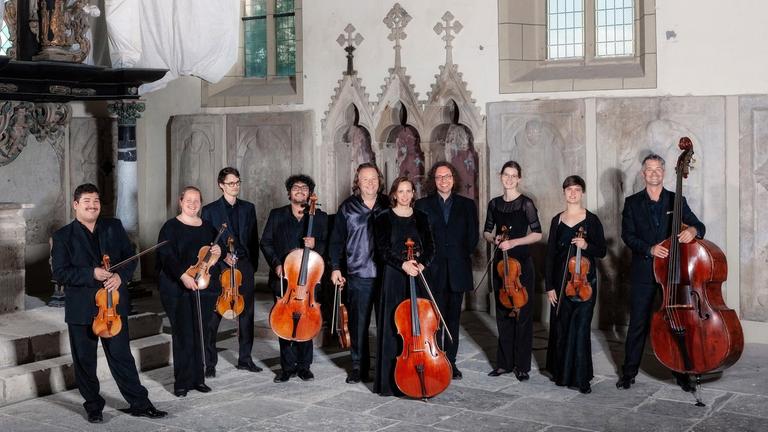Die Musiker des Ensembles stehen mit ihren Streichinstrumenten in einem hellen Kirchenraum. Die Musiker des Ensembles stehen mit ihren Streichinstrumenten in einem hellen Kirchenraum.