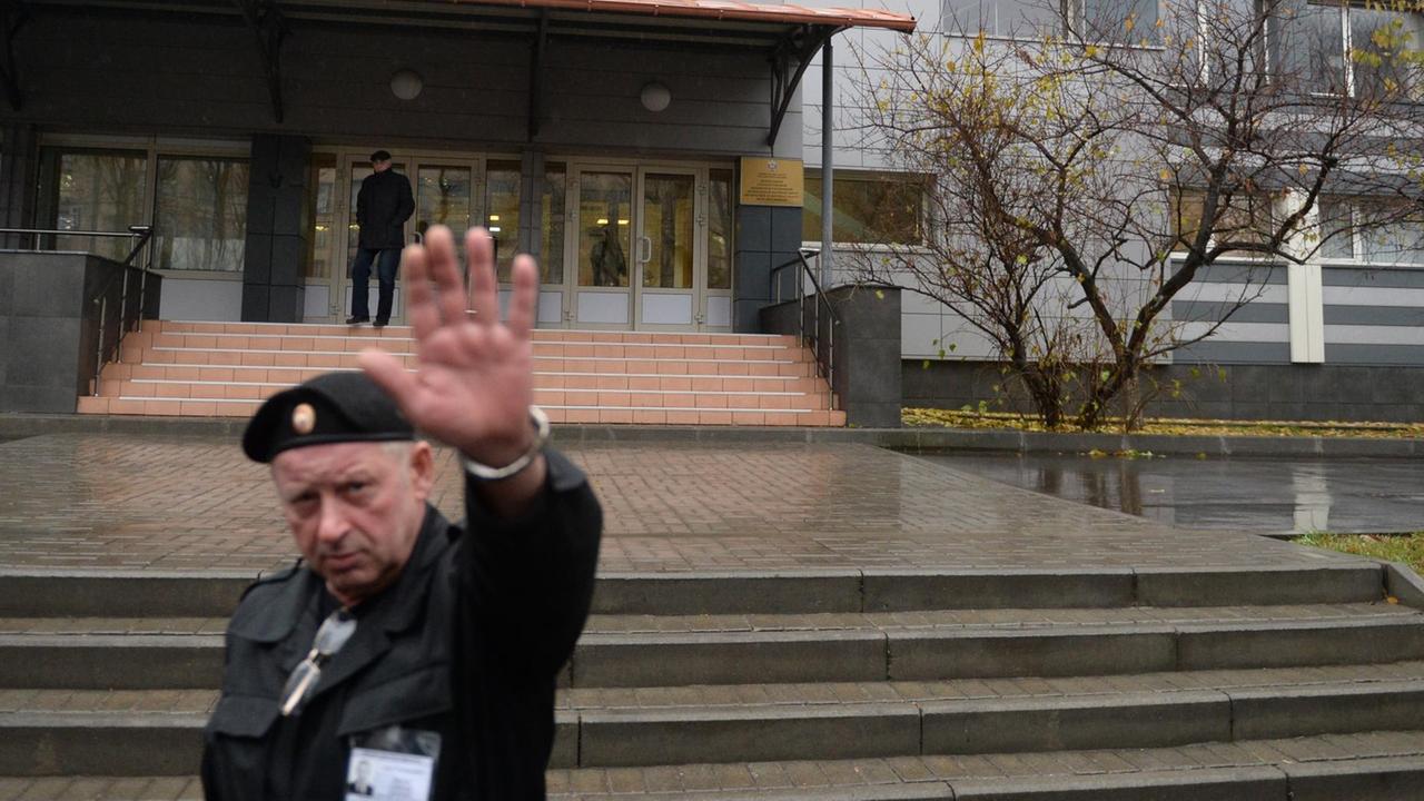 Der Wachmann mit Uniform und Barett hält vor dem Gebäude eine Hand in die Höhe. Die Treppe zum Eingang ist regennass. Der Wachmann mit Uniform und Barett hält vor dem Gebäude eine Hand in die Höhe. Die Treppe zum Eingang ist regennass.