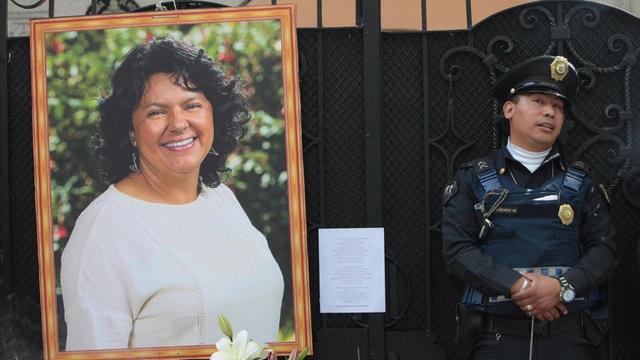 Ein Bild von Berta Cacéres an einem Zaun, daneben steht ein mexikanischer Polizist