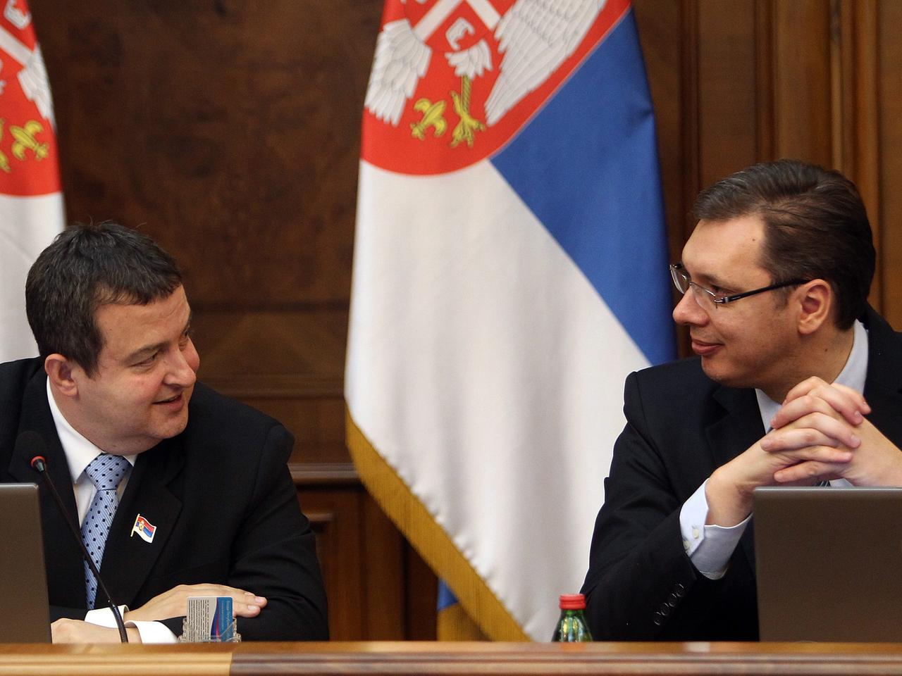 Ivica Dacic und Alexander Vucic sitzen an einem Tisch vor Laptops und unterhalten sich. Im Hintergrund vor einer branuen Holzwand hängen zwei serbische Flaggen in den Farben blau-weiß-rot.