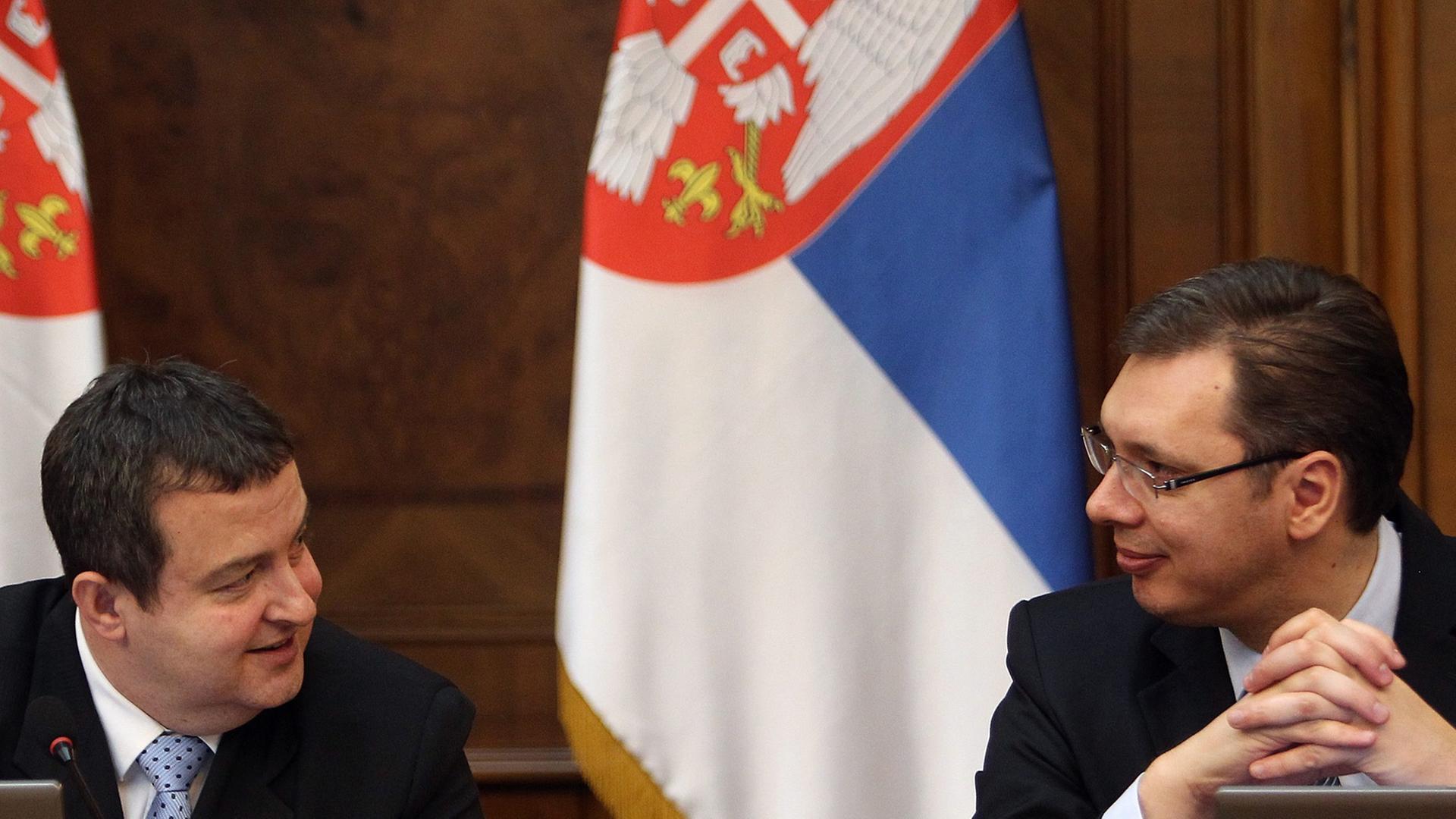 Ivica Dacic und Alexander Vucic sitzen an einem Tisch vor Laptops und unterhalten sich. Im Hintergrund vor einer branuen Holzwand hängen zwei serbische Flaggen in den Farben blau-weiß-rot. Ivica Dacic und Alexander Vucic sitzen an einem Tisch vor Laptops und unterhalten sich. Im Hintergrund vor einer branuen Holzwand hängen zwei serbische Flaggen in den Farben blau-weiß-rot.