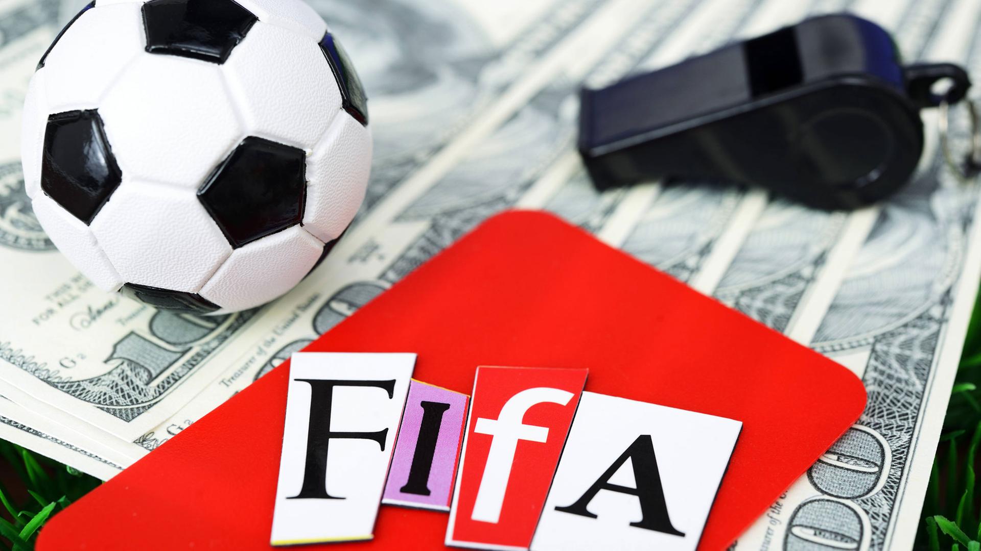 Ein Miniaturfußball auf Dollarscheinen neben einer schwarzen Trillerpfeife und einer Roten Karte mit Fifa-Schriftzug.
