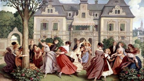 Festlich gekleidete, junge Menschen tanzen wirbelnd vor einer Schlosskulisse auf einem Rasen zwischen blühenden Büschen.