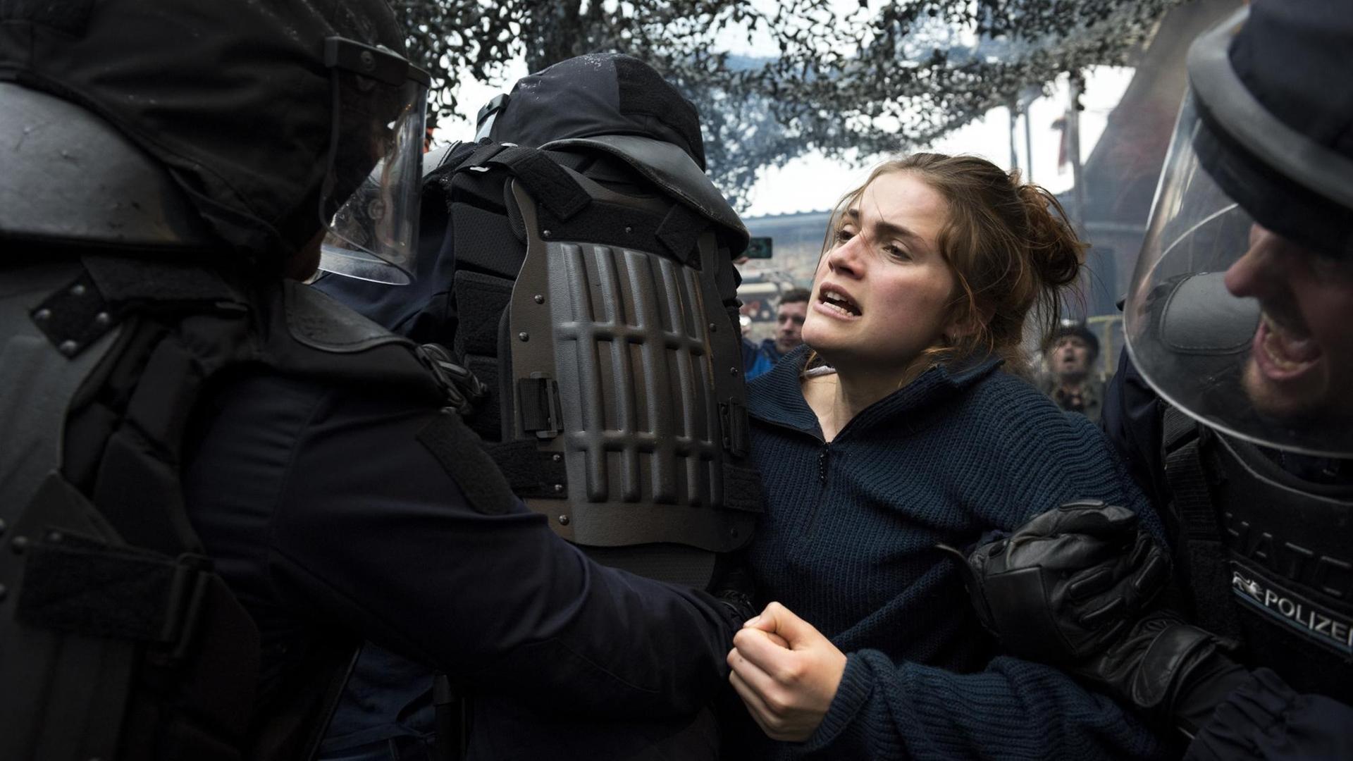 Eine junge Frau wird von mehreren vermummten Polizisten bedrängt und festgehalten. Sie hat ein ernstes Gesicht. Eine junge Frau wird von mehreren vermummten Polizisten bedrängt und festgehalten. Sie hat ein ernstes Gesicht.